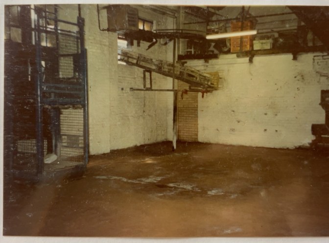 Original archive photograph: inside industrial premises in Croydon, showing a bare brick wall painted white with a blocked-up opening, a conveyor running overhead and a steel-caged goods lift to the left
